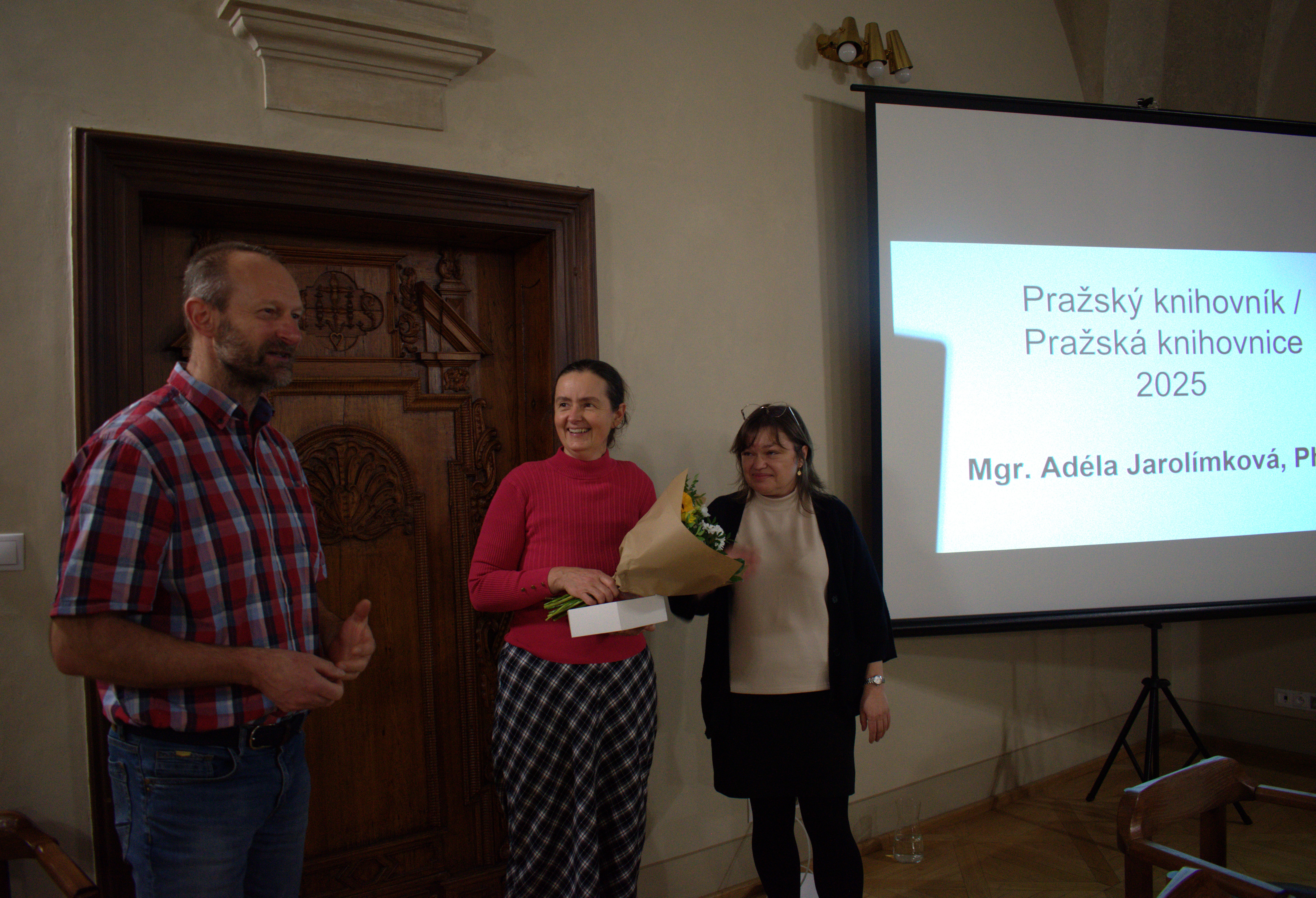 Žena  drží v ruce drží kytici, stojí mezi ženou a mužem, dívá se s úsměvem na muže, který hovoří k předpokládanému publiku. Vedle skupiny je promítací plátno, na kterém je zobrazen nápis Pražská knihovnice roku 2025 Adéla Jarolímková